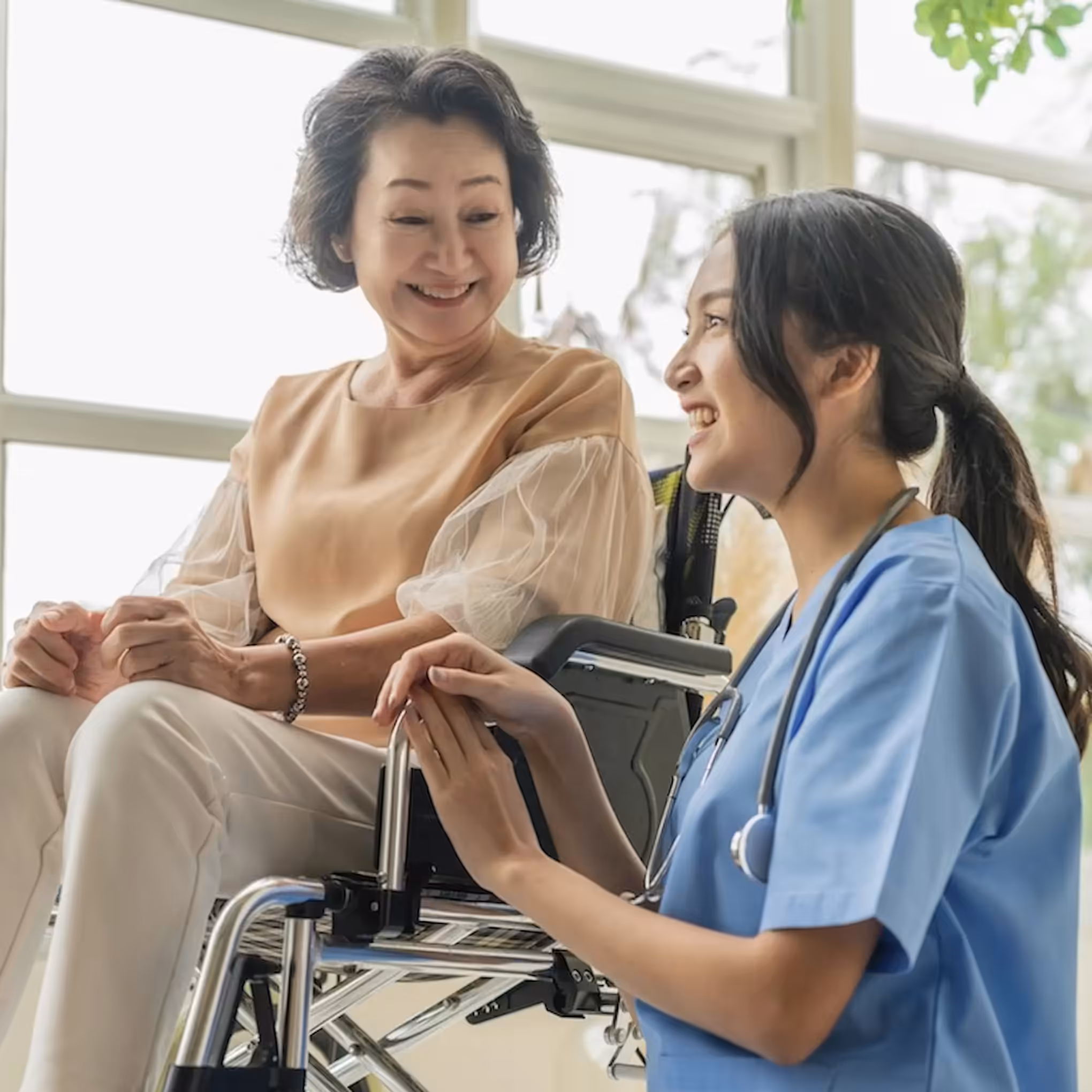 A smiling elderly woman sits in a wheelchair while a young female healthcare worker in scrubs kneels beside her, holding her hand and talking to her in a bright room, reflecting the warmth and compassion of quality home care.