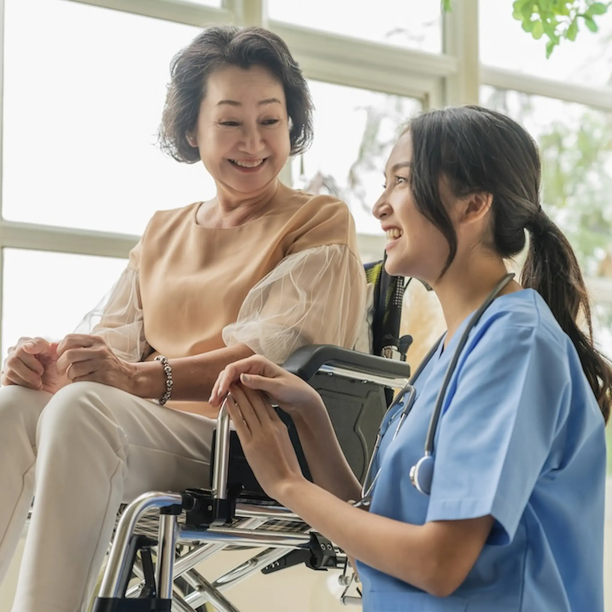 A smiling elderly woman sits in a wheelchair while a young female healthcare worker in scrubs kneels beside her, holding her hand and talking to her in a bright room, reflecting the warmth and compassion of quality home care.