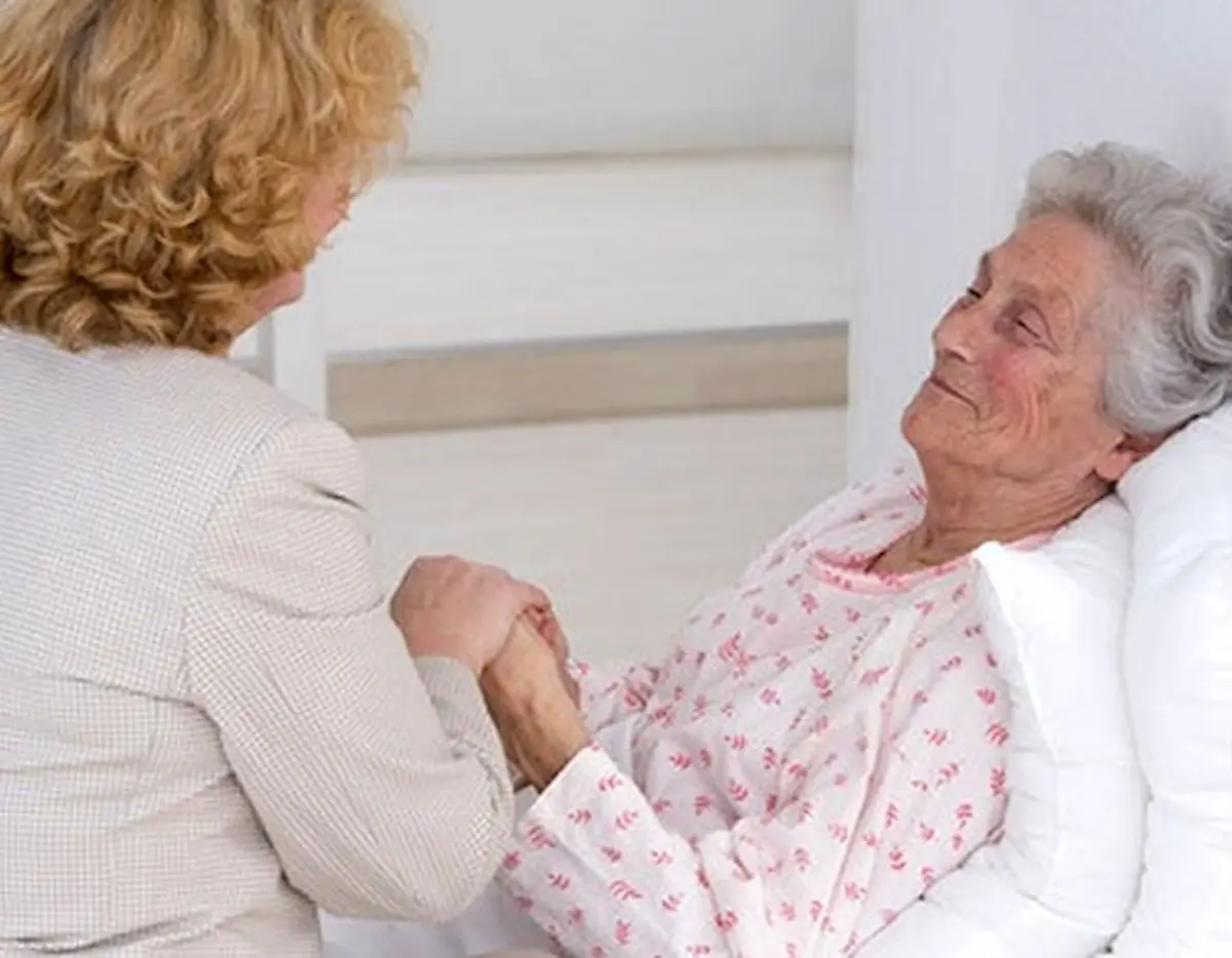 An elderly woman lies in bed, propped up by pillows, smiling and holding hands with another woman who is standing beside her, offering comfort and support.