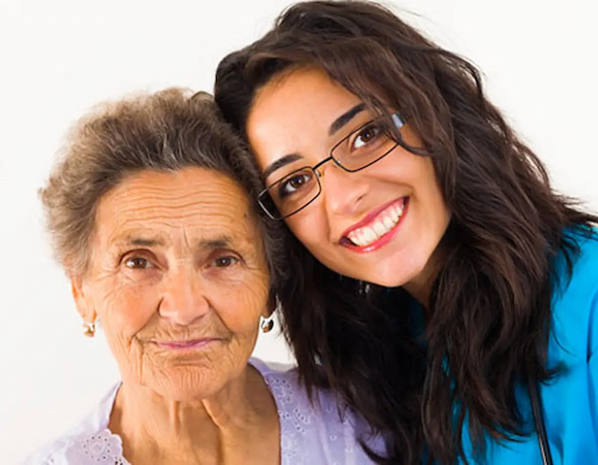 A smiling young woman in glasses and blue scrubs hugs an elderly woman with gray hair and a gentle expression; both women are close together against a light background.