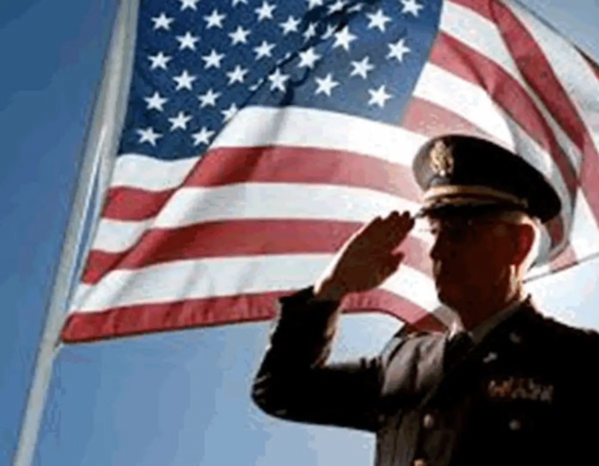 A uniformed military officer salutes in front of a large American flag waving in the background under a clear blue sky.