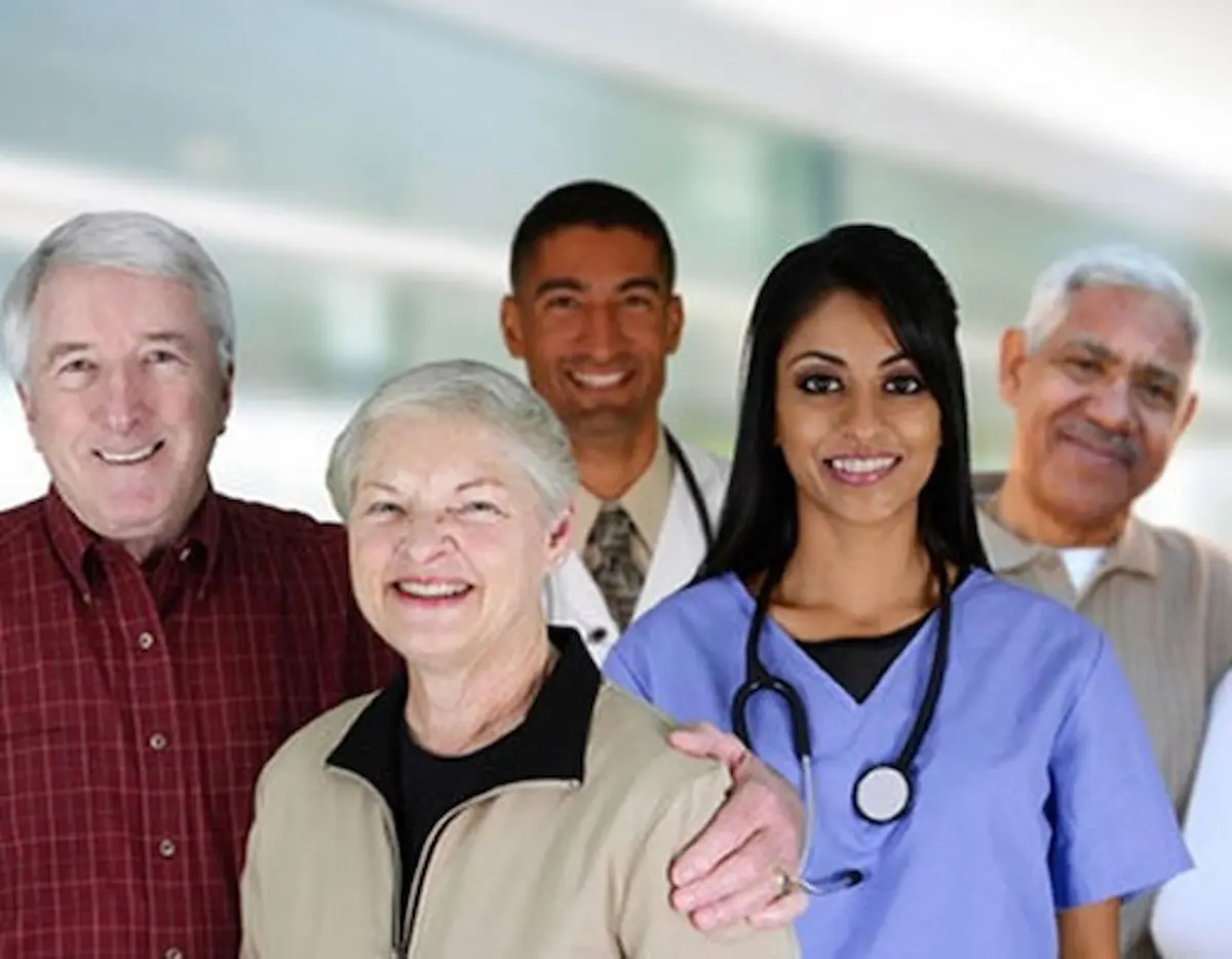 A group of smiling people, including two seniors in front and three adults behind them, one of whom is a healthcare worker in blue scrubs with a stethoscope. They stand in a bright indoor setting.